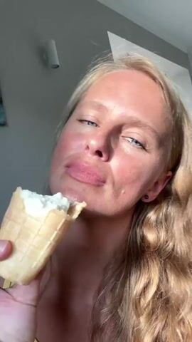 A blonde woman with blue eyes and rosy cheeks is eating ice cream from a waffle cone. She is holding the cone in her right hand and bringing it to her mouth. The background is blurry and appears to be indoors with sunlight coming from the left.