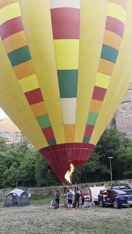 A large yellow hot air balloon with red, yellow, and green diamond patterns is being inflated on a grassy field. The burner is firing, indicating the balloon is actively being prepared for flight. Several people are gathered around the base of the balloon, appearing to be ground crew or passengers. Two tents and a blue SUV are visible in the background, along with a rocky cliff face and trees. A dog is sitting in the grass in the foreground. The sky is clear and blue.