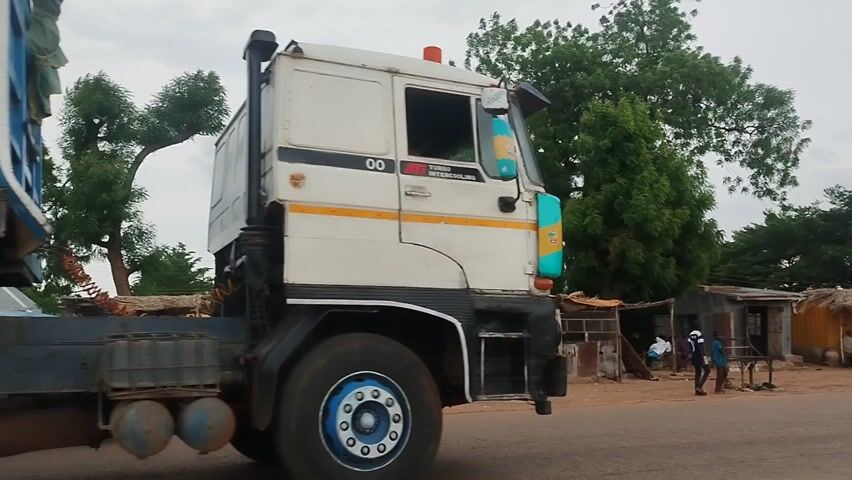 A white truck with a blue, open-sided trailer drives along a paved road from right to left under an overcast sky. The truck's cabin has "TURBO INTERCOOLER" and "00" written on its side. The trailer, initially appearing as a solid blue cargo box, is revealed to be a large, slatted structure, possibly for transporting livestock or goods, as the truck moves past the camera.

The truck's mudflaps at the rear of the trailer have the "DAF" logo, and a small white rectangle on the rear of the trailer displays "60-06". There are also reflective safety markings on the truck and trailer. In the background, another large truck covered with a tarp is visible further down the road.

The roadside features simple, corrugated metal structures or shacks and green trees. A person is briefly visible near one of these structures, and another individual appears to be walking in the distance. The road has dirt shoulders, and streetlights are visible further along the path. The video captures the truck's slow movement as it continues down the road in what appears to be a semi-urban or rural setting.