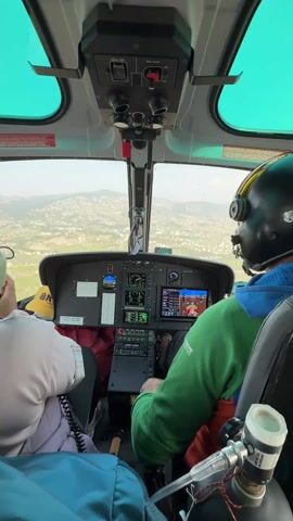 The video shows the interior of a helicopter cockpit during daytime, offering a pilot's perspective. Two individuals are seated inside; on the right, a person in a green jacket and black flight helmet, presumably the pilot, operates the controls. To their left, a passenger in a light-colored top is visible.

The cockpit features a complex control panel with multiple screens displaying flight information and what appears to be a navigation or terrain map. An overhead panel has switches labeled "TREAU TEST" and "ENG START OFF." Through the large windshield, a panoramic view of a hilly, semi-urban landscape is visible, with numerous buildings and green patches spread across the terrain, consistent with the Changunarayan, Nepal location.

Next to the pilot, an oxygen bottle is secured, with visible text reading "USE NO OIL OR GREASE ON VALVES" and "MEDICAL OXYGEN USP," indicating a safety or medical supply. A yellow object with "SKYLARK" written on it is partially visible near the passenger. The helicopter appears to be in flight, offering an elevated view of the surrounding area.