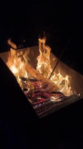 The video shows a close-up view of a square metal fire pit at night. Inside the fire pit, logs are burning with bright orange and yellow flames, and sparks are flying upwards. A person's hand is visible on the right side of the frame, holding a long, thin stick or skewer, which is inserted into the fire. The person appears to be poking or stirring the fire with the stick. The background is dark, indicating nighttime. The overall scene depicts a campfire experience.