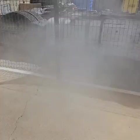 The video shows a tire burning behind a black chain-link fence. Smoke is billowing from the tire and obscuring the background. The ground in front of the fence is paved with a light-colored material, possibly concrete. There are some discarded tires and other debris visible in the background, as well as a building structure. The lighting suggests it is daytime.