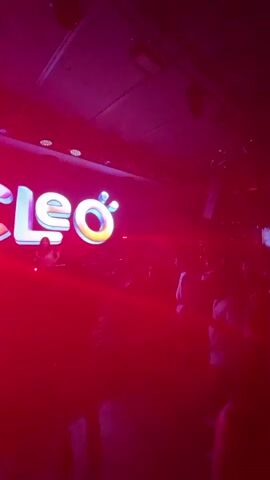 The video shows a dimly lit nightclub with a large illuminated sign that reads "CLEO". A DJ, a woman in a pink top, is visible behind a console on a raised platform under the sign. Red laser lights are cutting through the dark atmosphere. A crowd of people are standing and dancing in front of the DJ. Some individuals are silhouetted against the light. The overall environment suggests a lively party or club scene.