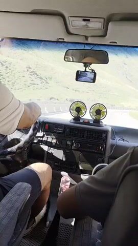 The video shows the interior of a car driving on a winding road through a mountainous landscape. The camera is positioned in the front passenger seat, looking forward. The dashboard is visible, with two small electric fans mounted on top of it. A smartphone is mounted above the rearview mirror, displaying a video feed. A driver's hand is visible on the steering wheel. The scenery outside the car consists of rolling green hills and a clear blue sky. The video appears to be a first-person perspective from inside the car.