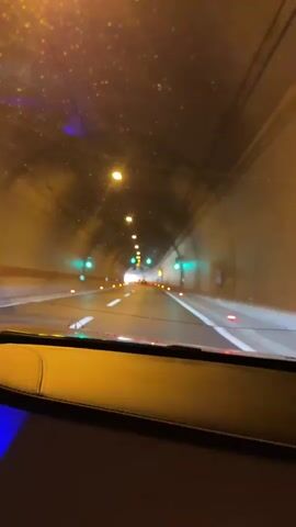 The video shows the view from inside a vehicle driving through a tunnel. The tunnel is illuminated by overhead lights and green traffic lights are visible on the sides. The road surface is visible, with white lane markings and red lights marking the sides of the road. The interior of the vehicle is partially visible, with a leather dashboard and blue ambient lighting. The tunnel walls are made of concrete. The overall impression is of a modern tunnel with good visibility.