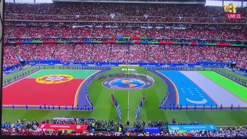 The media file shows an overhead view of a large, packed football stadium during an opening ceremony. Two national flags, Portugal and Uzbekistan, are prominently displayed on the pitch. A line of individuals, likely athletes or officials, stands on the field between the flags. In the center of the pitch, a circular logo with images of the Earth and the words "FIFA WORLD CUP RUSSIA" is visible. The stands are filled with spectators, most of whom are wearing red. The overall atmosphere appears to be celebratory and formal, typical of a major sporting event. The ZOR TV logo, along with the text "LIVE UZB RUS", is visible in the top right corner of the screen, indicating the broadcast is live and potentially from Uzbekistan or Russia. The time of day appears to be daytime, with the stadium well-lit. The weather is not discernible from the footage. The scene is set in a modern football stadium, likely in Mashrab, Uzbekistan, given the presence of the Uzbek flag and the "UZB RUS" broadcast information.