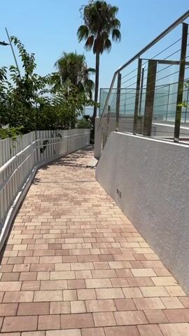 The video pans down a pathway paved with rectangular, tan and pink bricks. The pathway curves to the left and is lined on the left by a white fence with metal railings. Behind the fence are green trees and foliage. On the right side of the pathway is a white textured wall and a metal railing with horizontal bars. Behind the railing, a glass barrier is visible. Further up the pathway, palm trees are visible against a clear blue sky. The scene appears to be outdoors on a sunny day.