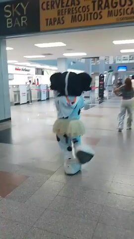 A person in a black and white spotted mascot costume with a light blue hoodie, a beige tutu, and white shoes is dancing with their back to the camera, performing hip movements. Their head is black with what appears to be a white stripe. In the background, to the right, a man in a white and green outfit stands holding a long green flag or banner. The scene takes place in what appears to be an indoor airport terminal with various counters and other people in the distance.