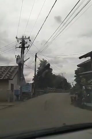 The video shows a view from inside a vehicle looking out at a road. The road curves uphill and is lined with trees and buildings on both sides. Overhead, numerous electrical wires crisscross the sky, supported by wooden poles. On the left side of the road, there is a house with a corrugated metal roof. On the right side, there are some buildings that appear to be shops. A motorcycle is seen approaching from the distance, going uphill. The sky is overcast.
