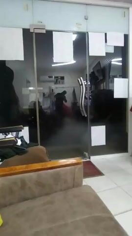 A video shows the interior of what appears to be a waiting area, with a brown upholstered couch in the foreground.  The view is through a partially tinted glass door leading into a barber shop or salon. White squares are affixed to the door. Inside, a person is indistinctly visible moving near a red barber chair.  Equipment is visible on shelves. The floor is tiled.