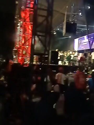 The video shows a live music performance at an outdoor venue. A band is on stage, with several members dressed in green uniforms. Behind them, a large screen displays the words "FA&M GRUPO." A crowd of people is gathered in front of the stage, some wearing red shirts. The stage is lit with bright lights, including green spotlights. The overall atmosphere appears to be lively and energetic.