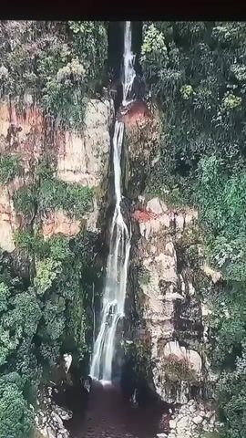 The video shows a drone's perspective of a tall waterfall cascading down a rocky cliff face covered in lush green vegetation. The waterfall originates from a narrow opening at the top of the cliff and descends into a dark pool of water at the bottom. The camera then pans to reveal the entrance of a large cave, also surrounded by dense foliage. Several people are visible standing near the cave entrance, appearing to be on a tour or excursion. The cave entrance is large and dark, with a visible rocky formation inside. The overall scene is one of natural beauty and exploration.