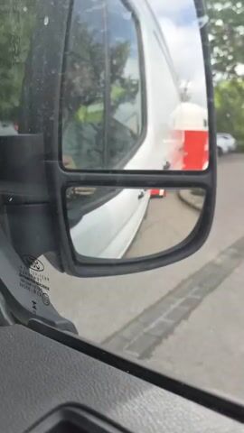 The view from inside a vehicle shows the side mirror.  The mirror reflects a white van in front of the vehicle and a red and white trailer attached to the van. The vehicle is stationary on a paved road.  There are trees in the background.  The vehicle's dashboard is visible.