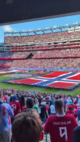 The video shows a stadium filled with spectators. In the center of the field, a large flag is being held open by people. The flag is red with a blue cross outlined in white. The stands are packed with people, many wearing red. Some people in the foreground are wearing red t-shirts, one of which has the name "BRAUT HAALAND" and the number 9 printed on it. The sky is blue and the weather appears to be sunny. A large screen at the top of the stadium displays an image related to sports.