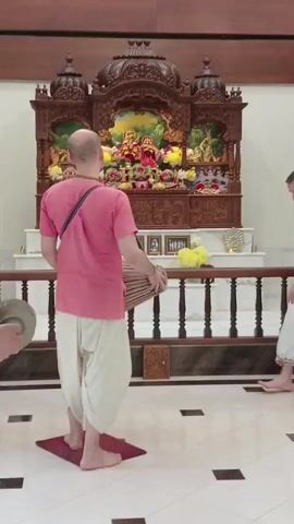 Here is a description of the video.

The video shows a group of people in what appears to be a temple or religious building. Several people, mostly men, are facing an ornate wooden altar decorated with flowers, statues, and religious imagery. A man in a pink shirt is playing a drum while standing on a small red rug.  The people are dressed in loose-fitting clothes, and some are bald. The floor is tiled in a white and black pattern. The room is well-lit, and curtains are visible in the background. One woman wearing a dark skirt is present.