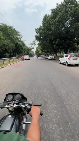 The video shows a first-person perspective of someone riding a black motorcycle on a paved road. The road is lined with trees on both sides. Several cars and auto-rickshaws are visible on the road, as well as some pedestrians. The sky is overcast. The rider's hand is visible on the handlebars.