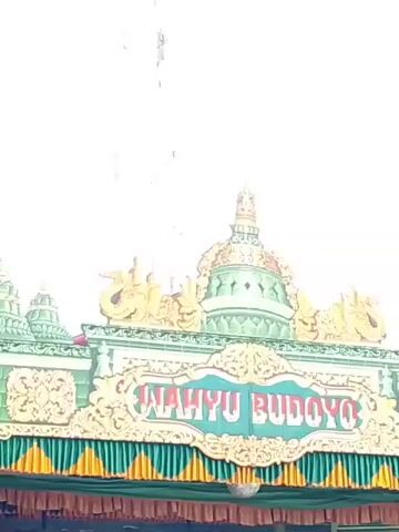 Here is a brief description of the video: 

The video shows a sign that reads "Wahyu Budoyo." The sign is green with gold accents and has a traditional design. There is a row of green and gold tassels hanging below the sign. The video is taken from a low angle, so the background is mostly white.