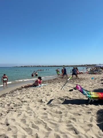 The video shows a crowded beach on a clear, sunny day. The sand is light-colored and wet near the water's edge. Several people are in the shallow water, swimming, playing, and wading. Some are further out in the sea, including one person on an inflatable. On the beach, there are people sunbathing, sitting on towels, and walking. A beach chair with a colorful pattern is visible in the foreground. In the distance, there is a faint outline of buildings along the coastline, and a small island with a structure on it can be seen in the sea. The sky is a bright, cloudless blue.