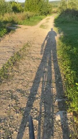 This video shows a person's long shadow stretching across a dirt road. The road is flanked by green grass and trees. The sky is clear and blue. The sun is setting, casting long shadows. The person is not visible in the frame, only their shadow. The video appears to be shot from the perspective of someone riding a bicycle or walking on the road.