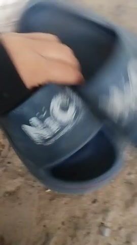 This media file features a close-up, overhead view of a person's hands interacting with several pairs of shoes on a sandy or dusty ground. The scene appears to be outdoors, possibly in a yard or a natural setting, given the texture of the ground.

The primary objects are two pairs of footwear:
1.  A pair of blue slides with a prominent white logo that reads "NIC NEW SHOES". The slides have a wide strap across the foot.
2.  A pair of white sneakers, which appear to be Nike Air Force 1s, identifiable by their distinctive sole design and the Nike swoosh logo.

The activity captured is the person picking up and handling the shoes. The motion blur suggests quick movements, indicating the person is either showing off the shoes, organizing them, or perhaps preparing to put them on or put them away. The lighting suggests it might be daytime, but no specific time can be determined. There are no clear indicators of weather conditions or a specific time of day beyond daylight.

The hands are the only visible human element, and their skin tone suggests a person of average complexion. No emotions are discernible from the brief interaction shown. The setting is unadorned, focusing solely on the shoes and the ground.

There is no indication of the location being Cîrpești, Moldova within the media file itself. This information is external to the visual content.