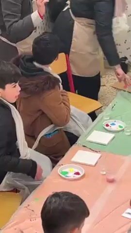 The video shows a group of people, primarily children, participating in what appears to be an art activity. They are seated around tables covered with pink and green plastic tablecloths. Each person has a white canvas in front of them along with small dishes containing paints in various colors. Most individuals are wearing aprons. The instructor is visible, standing and gesturing while facing the group. The room seems to be indoors, likely a classroom or studio setting. The overall atmosphere appears to be focused on creativity and learning.