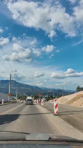 The video shows a first-person perspective of driving on a road under construction. The road is paved, and there are temporary traffic cones and barriers in place. Several cars are visible on the road, moving in the same direction as the camera. In the background, there are mountains and a bright blue sky with white clouds. There are also power lines visible overhead. The overall impression is a typical scene of roadwork on a sunny day.