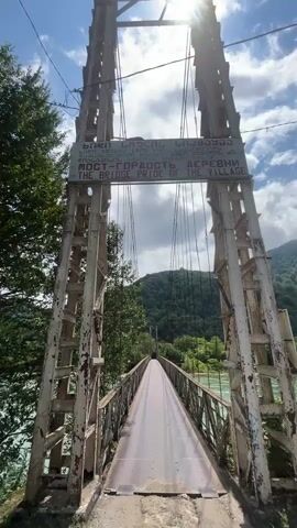 The video opens with a low-angle, first-person perspective shot of a suspension bridge. The bridge appears old and weathered, with rust visible on the metal structure. Two tall, ornate support towers frame the entrance to the bridge, each adorned with a sign. The sign at the top of the bridge is multilingual, displaying text in what appears to be Georgian, Russian, and English, with the phrase "THE BRIDGE PRIDE OF THE VILLAGE" clearly visible. The bridge deck is made of metal plates and slopes downwards into the distance. To the left of the bridge, lush green trees and foliage are visible, and to the right, the turquoise waters of a river flow rapidly. The background is dominated by densely forested green hills under a bright, partly cloudy sky. The camera slowly moves forward, entering the bridge and continuing down its length. As the camera progresses, the river becomes more prominent on both sides, and the surrounding landscape of hills and trees fills the view. The perspective remains fixed, giving the impression of someone walking across the bridge. The video ends as the camera is about halfway across the bridge, with the distant end of the bridge and the far bank of the river in view.