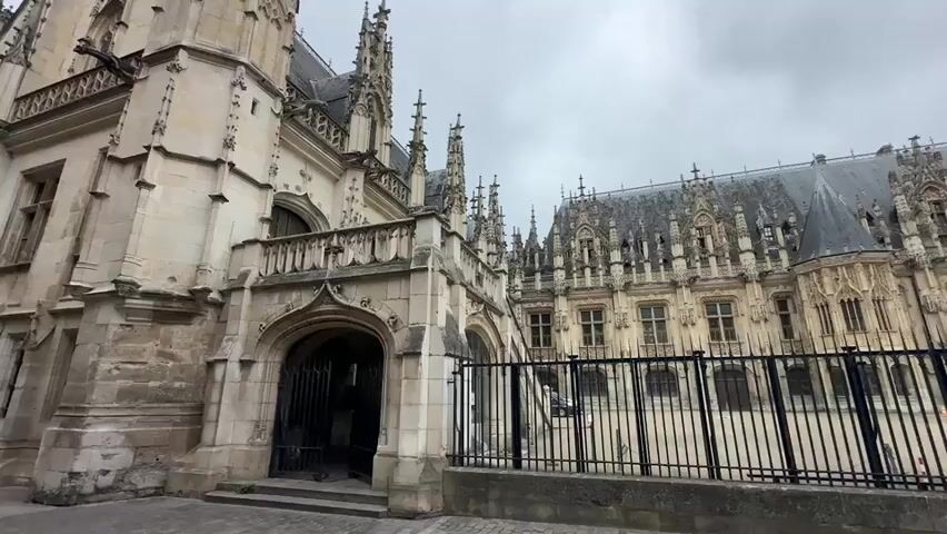 A wide shot captures the exterior of a large, ornate stone building with gothic architectural elements under a cloudy sky. The building features multiple spires, arched windows, and intricate stone carvings. A black metal fence runs along the front of the building, separating it from a paved walkway. The left side of the frame shows a close-up of a grand arched entranceway with a black metal gate. The right side of the frame shows more of the building's facade, with a French flag visible on the far right. The scene is static, with no visible people or activity. The lighting suggests an overcast day.