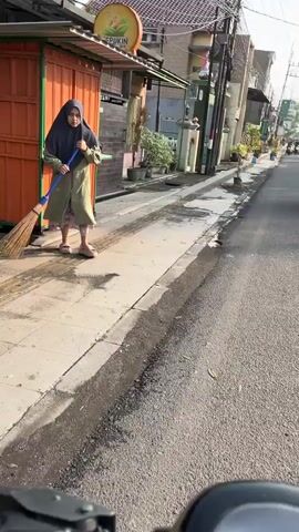 A person wearing a hijab and a long green coat is sweeping the sidewalk with a broom. The person is standing next to an orange building that has a sign with a picture of fruit. The sidewalk is made of concrete and is next to a street with asphalt. There are some potted plants along the sidewalk. The street appears to be in a residential or commercial area, with buildings on both sides. The lighting suggests it is daytime.