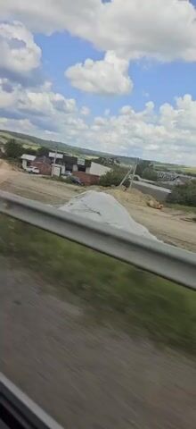 The video shows a view from a vehicle traveling on a road. The road appears to be rural or suburban, with buildings and construction materials visible on the side. A guardrail runs along the edge of the road in the foreground. The sky is blue with scattered white clouds. There are some traffic signs visible, including a yellow triangular sign with a black symbol that appears to be a warning. Several large concrete pipes are stacked on the ground. The overall impression is that of a typical roadside scene during daytime travel.