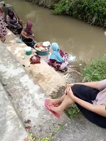 Four women are washing clothes in a shallow, muddy canal. One woman, wearing a red and pink patterned dress and a red headscarf, stands on a concrete ledge to the left, holding a large red basin filled with clothes. Another woman, in a dark blue and white patterned dress and a pink headscarf, is seated further down the ledge, also by the water. A third woman, in a floral print dress and a light pink headscarf, is crouched by the water's edge, actively washing clothes in a black bucket. A fourth woman, wearing a purple and white patterned dress and a light blue headscarf, is also crouched by the water, washing clothes in a white basin. There are other laundry basins and buckets scattered around them. The environment is lush with green vegetation on the opposite bank of the canal. A person's legs, wearing pink flip-flops, are visible in the foreground on the concrete steps.