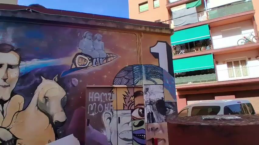 This is a video of a mural on the side of a building. The mural depicts a man riding a horse, with a space background. The man is wearing a white shirt and has a large head. The horse is brown and is wearing a saddle. In the background, there are stars and a planet. There is also a number 1 painted on the wall. The mural is colorful and detailed. The video also shows some of the surrounding environment, including a building with balconies, a street, and some trees.