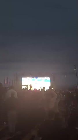 A crowd of people are gathered outdoors at night watching a large screen displaying a broadcast of a sporting event. The screen is brightly lit, and the surrounding area is dark, with some colored lights from the screen reflecting on the crowd. People are silhouetted against the light, and some appear to be cheering or raising their hands. The event seems to be happening in an urban environment, with a tall, modern building visible in the background to the left of the screen. The sky is dark and cloudy, suggesting it might be evening or nighttime.