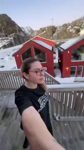 The video shows a woman standing on a wooden deck, holding the camera in selfie mode. She has long brown hair and is wearing glasses and a black t-shirt. Behind her are two small, red, A-frame houses with white trim. The ground is covered in snow. The camera pans to the right, showing the side of one of the houses and then revealing a view of a body of water with snow-covered islands in the distance. There are more red houses visible on the opposite side of the water. The camera then focuses on a round wooden table on the deck, which is also covered in snow. The sky is light blue and clear.