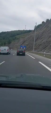 A forward-facing vehicle's dashboard is visible in the foreground. Outside the vehicle, on a highway, a white van travels in the left lane and a black Volkswagen Golf travels in the right lane. A blue road sign with white lettering and symbols is visible on the right side of the highway. The sign indicates a sharp curve to the left and a speed limit of 80. The highway is flanked by a steep, rocky slope on the right and a forested hill on the left. The sky is overcast.