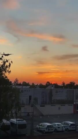 The video captures a serene sunset scene in Otopeni, Romania, as a large, four-engine turboprop aircraft, possibly a C-130 Hercules, flies across the colorful sky. The aircraft, silhouetted against the vibrant orange, yellow, and pink clouds, appears to be flying at a relatively low altitude, with what looks like exhaust plumes trailing from its engines.

In the foreground and middle ground, several buildings are visible, including one prominently displaying a large "HILS" sign with a phone number "0210 067 174" below it. Another sign with "KIM" partially visible can be seen further in the background to the right. Below the buildings, a parking lot contains several white cars and a bus or large van. Trees line the horizon, partially obscuring the setting sun, which casts a warm glow over the entire landscape. A tennis court is also visible on the right side of the frame, near the parking area. The sky transitions from a deep orange at the horizon to lighter hues of pink and blue higher up, indicating either sunrise or sunset.