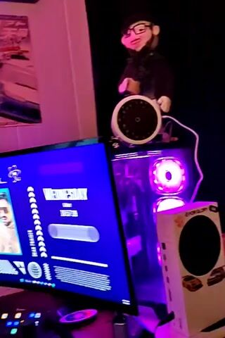 The video shows a computer setup in a room lit with pink and purple lights. Two monitors are visible, one displaying a customized desktop screen with widgets, including a clock and profile pictures, and the other displaying a fantasy-themed wallpaper with a figure standing before a large sword. Posters of cars decorate the wall above the monitors, and a small shelf is visible. A keyboard and a small figurine sit on the desk in front of the monitors. The video pans to the left.