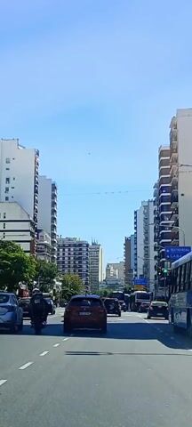 The video shows a street scene in an urban setting with tall buildings lining both sides of a multi-lane road. Cars and motorcycles are moving along the road, some of which appear to be stuck in traffic. The sky is clear and blue. There are street signs visible, with an arrow pointing straight and signs with text that is illegible at this resolution. The traffic light is green.