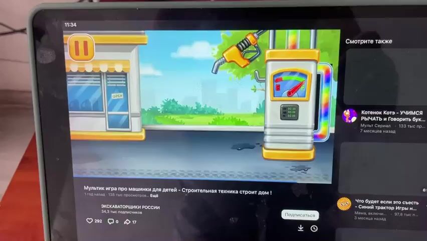 The video shows a tablet screen displaying a children's cartoon game. The game appears to be about construction vehicles building a house. The scene transitions from a gas station with a refueling nozzle to a partially built house, and then to a green truck. A pause button is visible in the top left corner of the screen. The video is paused at the 0:04 mark, showing the green truck approaching a red house with a gray roof and stone foundation. Text below the video indicates it is a cartoon game for children about construction technology building a house. To the right of the video, there are suggested videos displayed. The tablet is placed on a wooden surface.