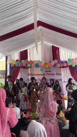 The video shows an awards ceremony taking place under a white tent with red trim. A stage is set up at the back with a banner that reads "Akhirussanah, Pentas Seni & Wisuda Tahfidz Al-Quran". Several young people, dressed formally and wearing sashes and medals, are standing on the stage. A woman in a traditional patterned dress is speaking into a microphone on stage. In the foreground, several people, mostly women wearing hijabs, are seated and watching the ceremony, some holding up phones to record. There are balloons above the stage and floral decorations. The event appears to be a graduation or recognition ceremony.