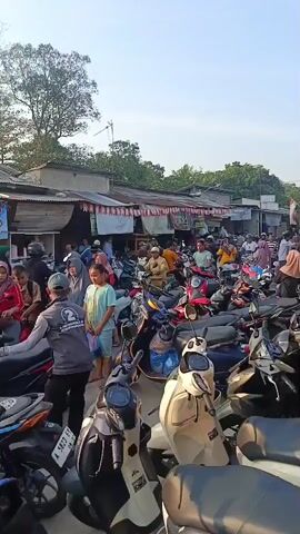 The video shows a crowded outdoor market scene, likely in a Southeast Asian country given the presence of many motorbikes and the attire of some individuals. The foreground is dominated by a densely packed parking area filled with numerous motorbikes, varying in color and style. Some motorbikes are parked closely together, suggesting limited space. Behind the motorbikes, a bustling market is visible. Several stalls with awnings and simple structures are lined up, displaying various goods, though specific items are not clearly identifiable. A large number of people are present, moving through the market and navigating the motorbike parking area. Many are dressed in casual clothing, with some women wearing headscarves (hijabs). One individual in the center, wearing a grey vest with the number "2" on the back, appears to be directing or interacting with others in the parking area. The overall atmosphere appears lively and busy, with people shopping and commuting. The lighting suggests it is daytime, possibly late afternoon, with the sun casting shadows. The environment appears somewhat informal, with basic construction of the market stalls.