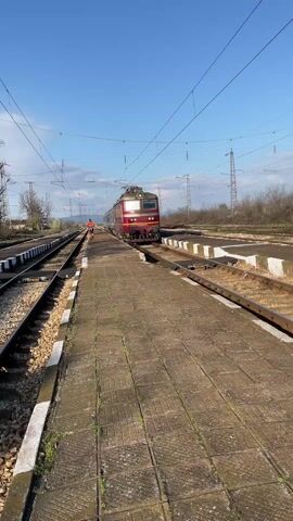 Here's a description of the video:

The video shows a red and gold passenger train moving past the camera on a train platform. The train is being pulled by a locomotive, and is made up of blue passenger cars with graffiti on the side. The platform is made of concrete blocks and has white-painted edges. An individual wearing a red shirt is standing by the side of the tracks, watching the train pass. Overhead are train cables and electrical poles against a bright blue sky. The setting looks like a rural train station.
