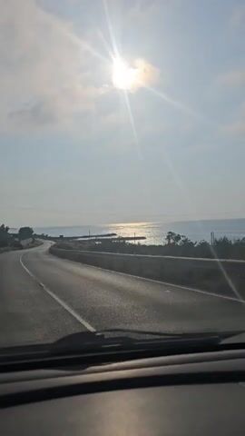 The video shows a view from inside a vehicle driving on a paved road that curves to the left. The road has white lane markings. To the right of the road is the sea, with sunlight glinting on the water. The sun is visible in the sky, appearing bright and partially obscured by clouds. There are some faint power lines visible in the upper part of the frame. The overall scene suggests a coastal drive on a sunny day.