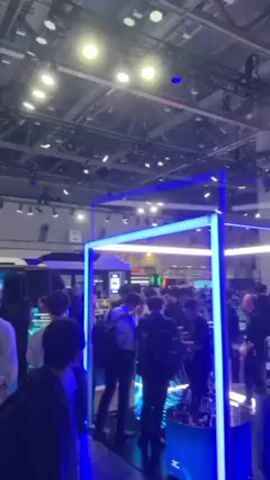 The video depicts a bustling scene at a large indoor event or trade show. There are many people walking around, mostly dressed in casual clothing, browsing various booths and displays. A prominent feature is a square structure outlined with bright blue neon lights, drawing attention to the area within it. Overhead, the ceiling is visible, revealing an extensive network of pipes, lighting fixtures, and other structural elements. Further back, a large screen displays a vibrant and colorful city scene at night. The overall atmosphere suggests a lively and modern technological exhibition.