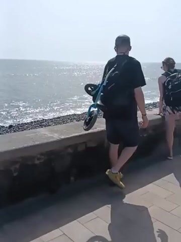 The video depicts a sunny, breezy seaside promenade. The camera follows a man carrying a folded blue stroller on his back and walking alongside a woman. They are walking on a brick-paved path that runs parallel to a low concrete wall bordering a beach. The sea is sparkling in the sunlight. The promenade is filled with people strolling, sitting on benches, or riding bicycles. Palm trees line the path, and there are buildings visible in the background.