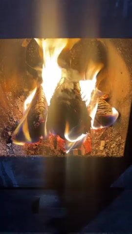 The video shows a close-up view of logs burning in a fireplace. The flames are bright yellow and orange, with some blue at the base. The logs are charred and glowing red in places. Ash and embers are visible at the bottom of the fireplace. The lighting is dim, with the light from the fire illuminating the scene.