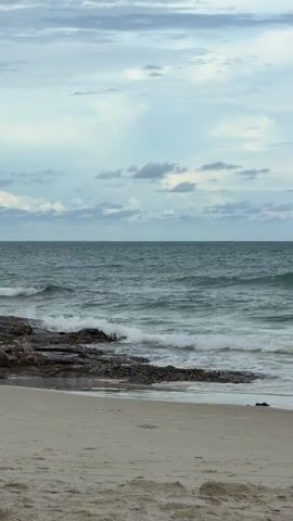 The video shows a natural scene of a beach with the ocean and sky. The foreground consists of sand, which appears to be a light beige color. In the middle ground, there is a rocky outcrop jutting out into the water. These rocks are dark and appear to be covered in some organic material, possibly seaweed or barnacles. The ocean is visible behind the rocks, with waves breaking and creating white foam along the shore. The water is a deep teal or greenish-blue color, and the waves suggest a moderate swell. The horizon line is clearly defined, separating the sea from the sky. The sky is overcast with a mix of light grey and blue clouds. The lighting suggests it is daytime, but not necessarily bright sunlight, possibly an overcast or cloudy day. There are no people or man-made objects visible in the frame. The overall impression is one of a natural, somewhat rugged coastline.
