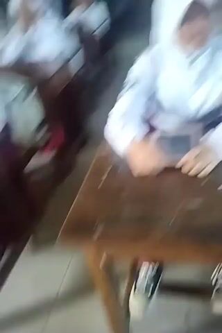 The video shows a classroom setting with students seated at desks. A teacher, wearing a red patterned shirt and a hijab, is standing and gesturing, seemingly addressing the students. Several students are wearing white shirts and headscarves, typical school uniforms. One student, a boy, is standing between the rows of desks, also appearing to be addressed by the teacher. Some students are looking at their devices, possibly tablets, placed on their desks. The classroom environment appears to be well-lit with natural light coming from windows. The overall scene suggests an educational activity or discussion taking place in a school.