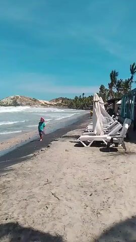 The video depicts a scenic beach scene on a bright, sunny day. The sky is a clear blue. Two people are walking along the shore near the water's edge. Waves are crashing onto the beach. A man is standing by himself at the water's edge. In the foreground, there are several white lounge chairs with folded white towels on them, lined up parallel to the beach. The sand appears to be a mixture of light and dark colors. In the distance, there is a small, rocky cliff or hill covered with vegetation. The overall ambiance is peaceful and relaxing.