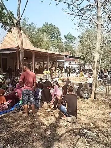 A group of people are gathered outdoors at what appears to be a ceremonial site. In the background, a building with a tiled roof and open-air covered walkways is visible. A group of individuals dressed in traditional yellow and patterned clothing are performing a dance or ritual on a raised platform in front of the building. Another group, dressed in dark clothing, is seated or standing behind the performers. In the foreground and middle ground, a crowd of onlookers is seated or standing on the ground, many of them wearing dark clothing, including headscarves. The ground is unpaved and appears to be dirt and dry leaves. The overall atmosphere suggests a cultural or religious gathering.